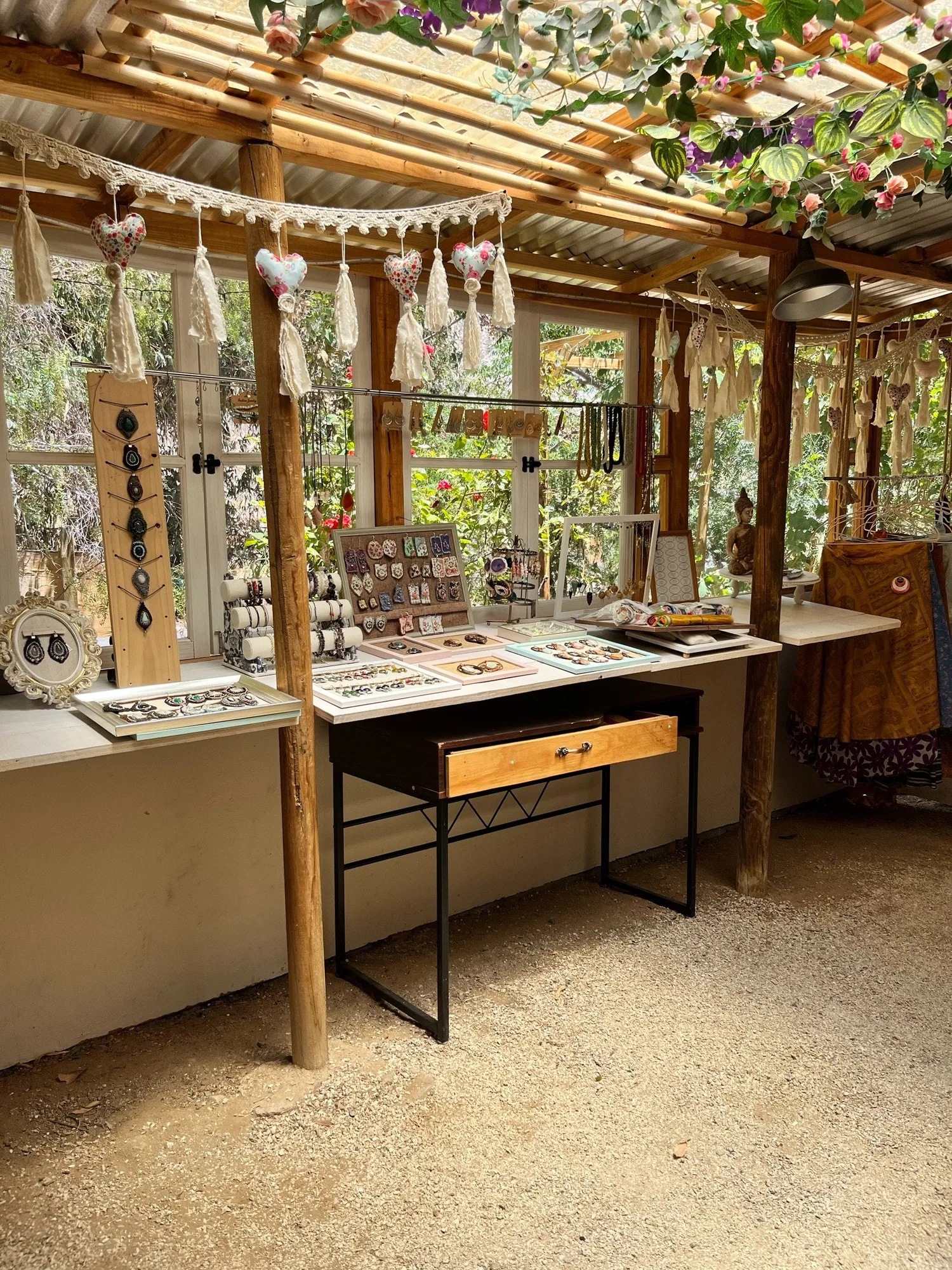 El mesón de la tienda con aros y collares hechos a mano, bajo guirnaldas de tela y la pérgola de caña